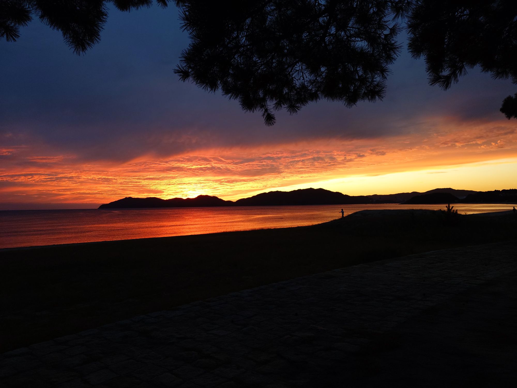 A shot over the sea of a brilliant red and orange sun setting over an island. There are clouds blotting out much of the sky.