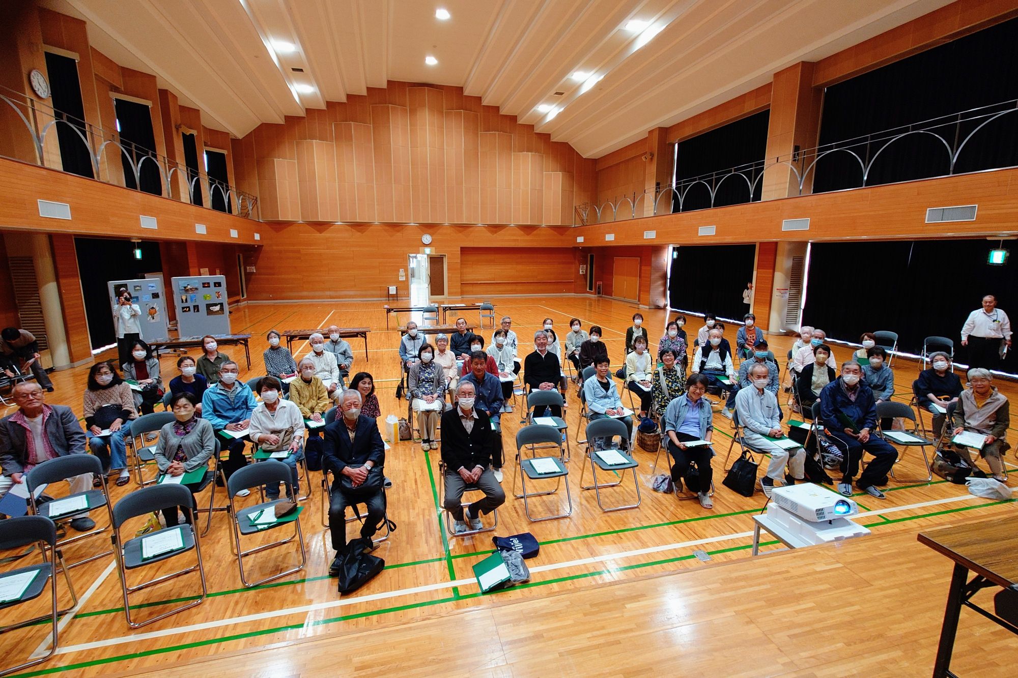 A small audience of older Japanese people sitting on folding chairs in a gymnasium-style hall.