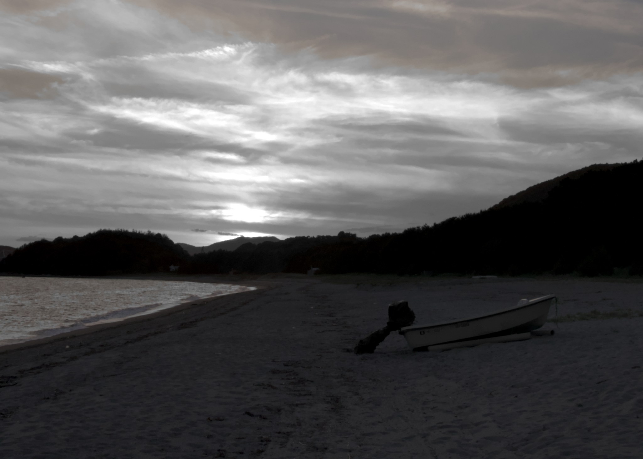 A B&W shot of a beach. The sun is setting behind a layer of clouds. In the foreground, a small motorboat is pulled up away from the water.