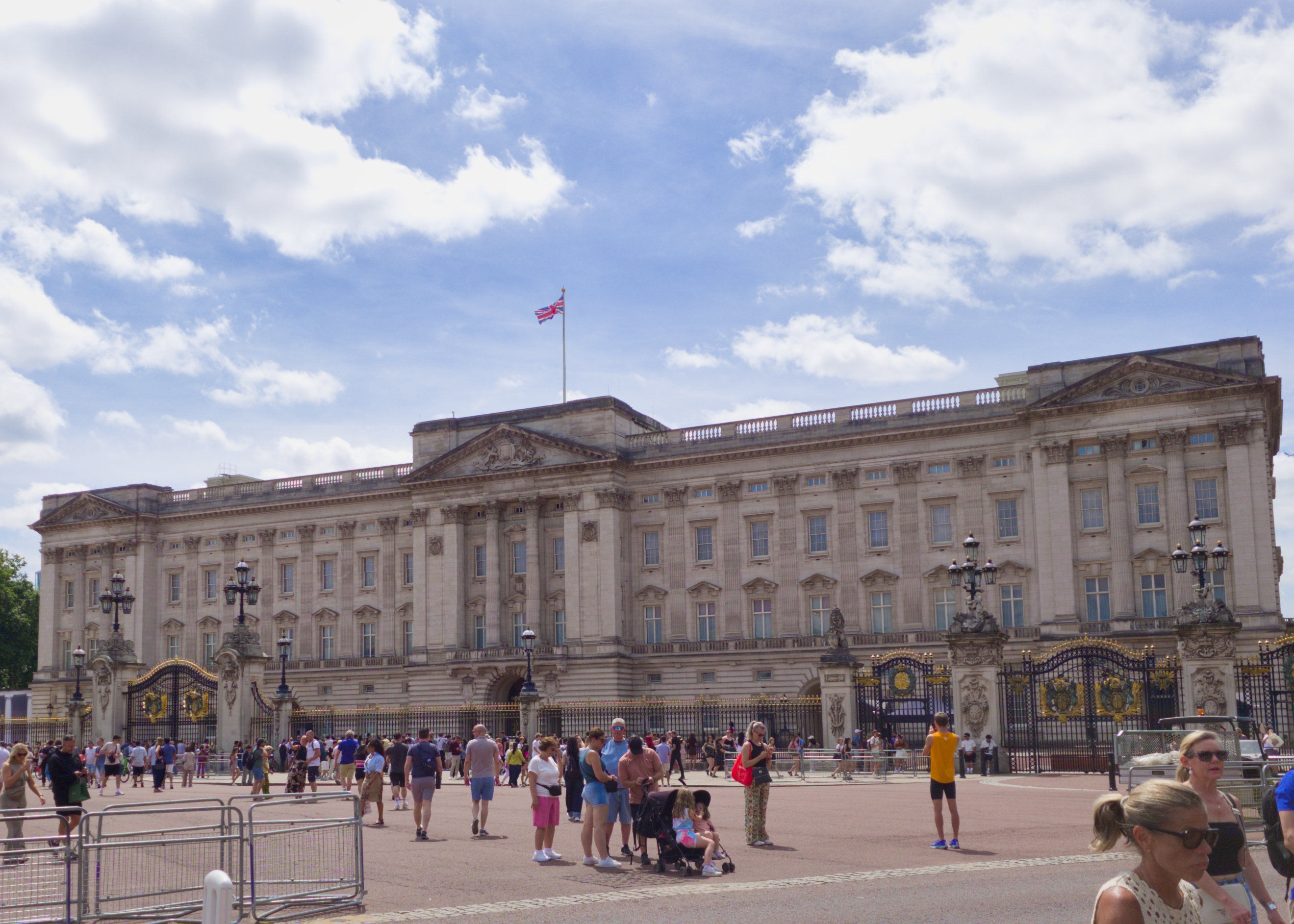 A wide shot of Buckingham Palace in London. The sky is blue, the sun is bright, and crowds of people stand in front of it.