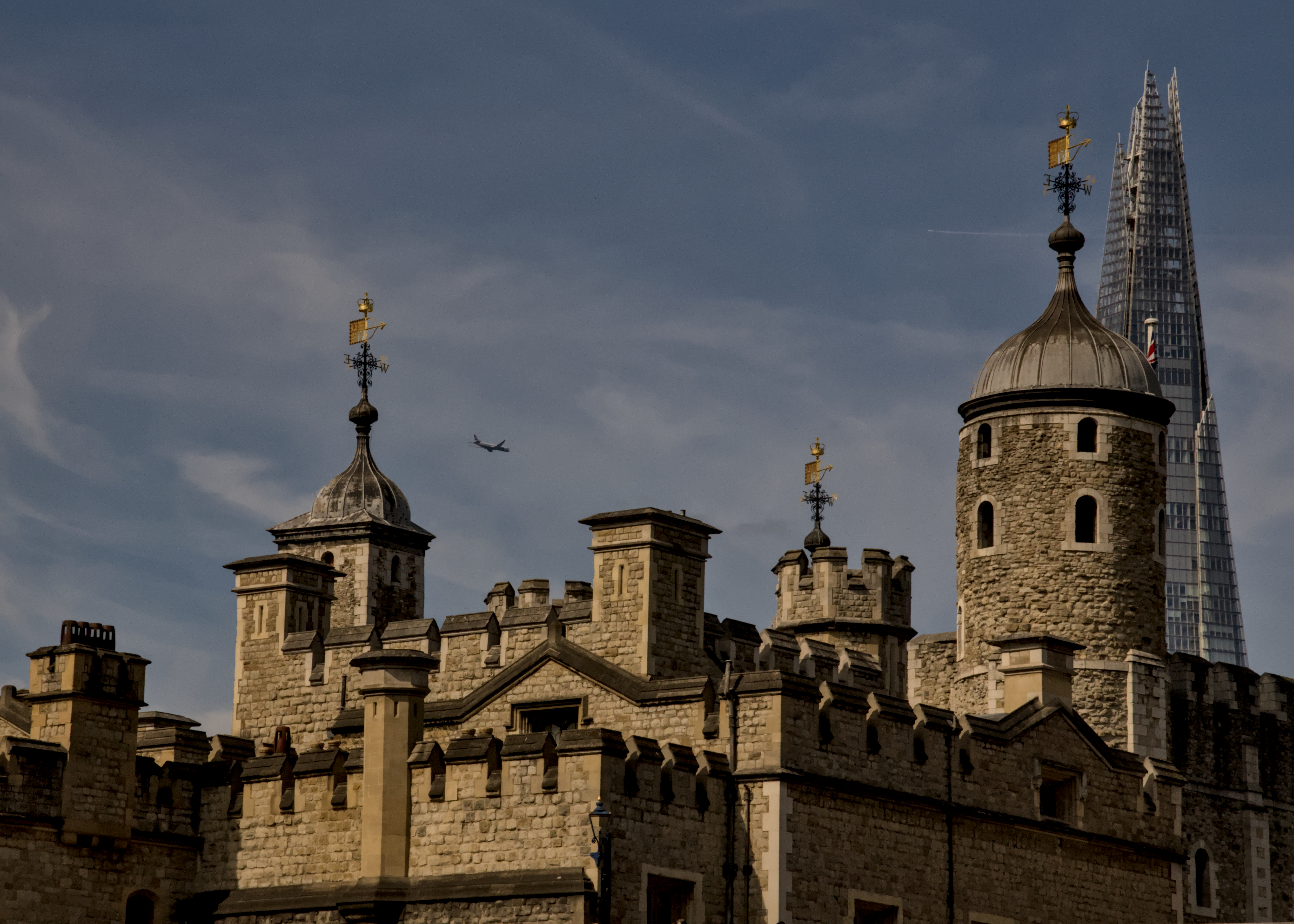 A view across the medieval looking crenellations and towers of the fortress known as the Tower of London. In the background is a pointed glass tower of a modern building, and a plan flies across the sky.