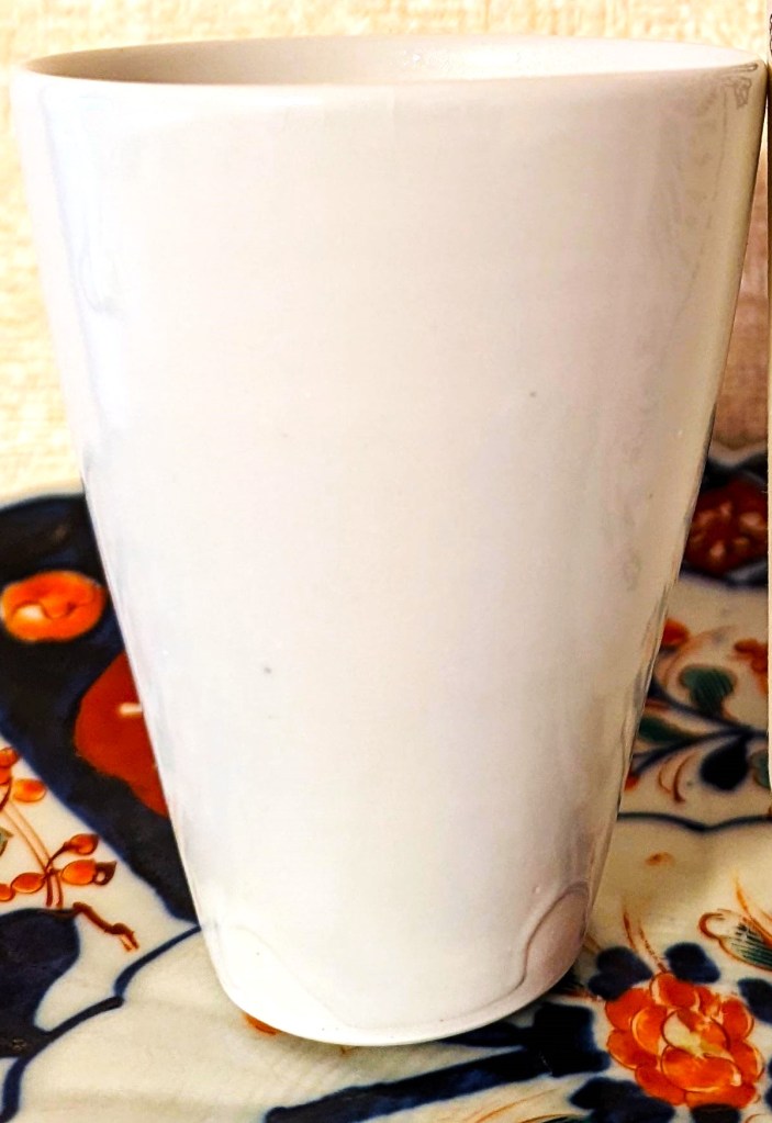 A tall white porcelain cup that narrows at the bottom. The glaze shows a finger-shaped open patch at the bottom.