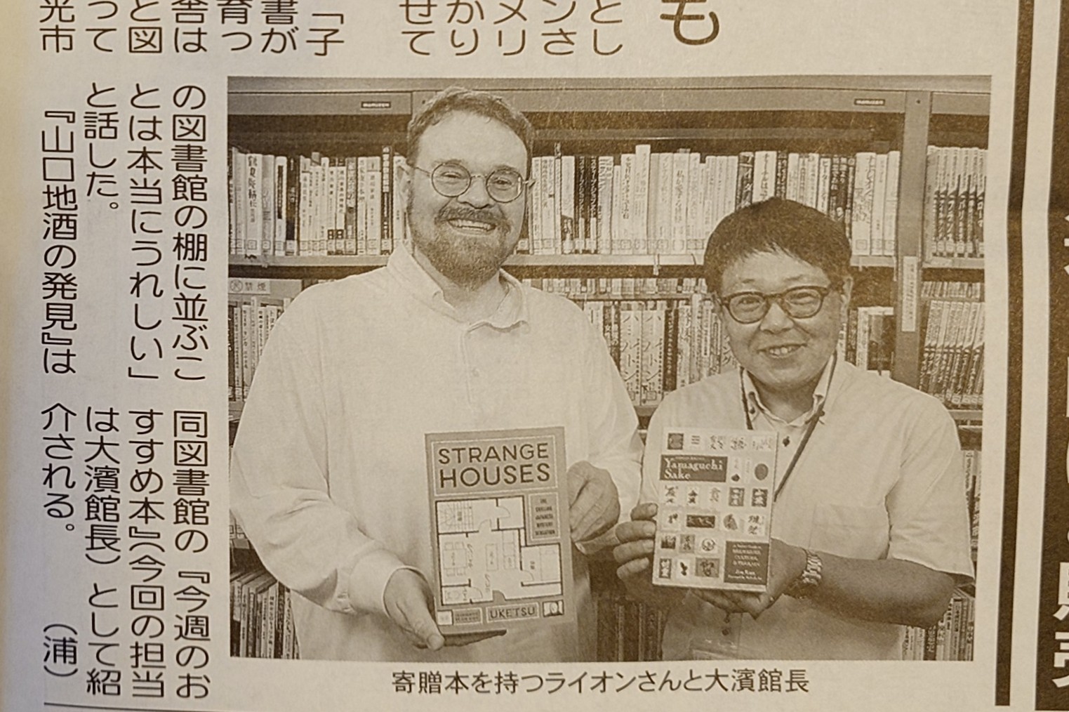Two men in glasses standing in front of a bookshelf holding two books, smiling at the camera.